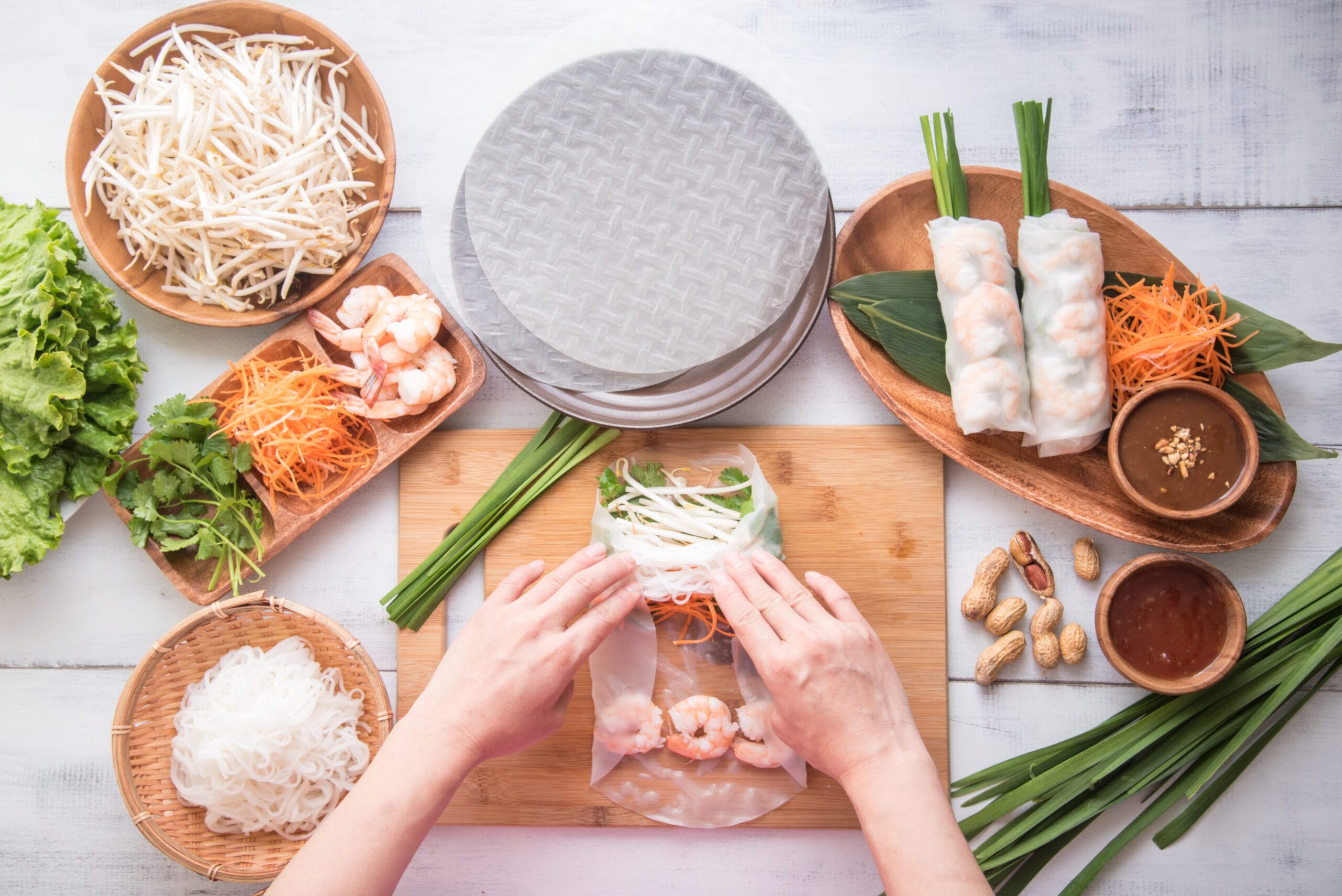 hands getting ready to roll the spring rolls; ingredients are scattered around the frame.
