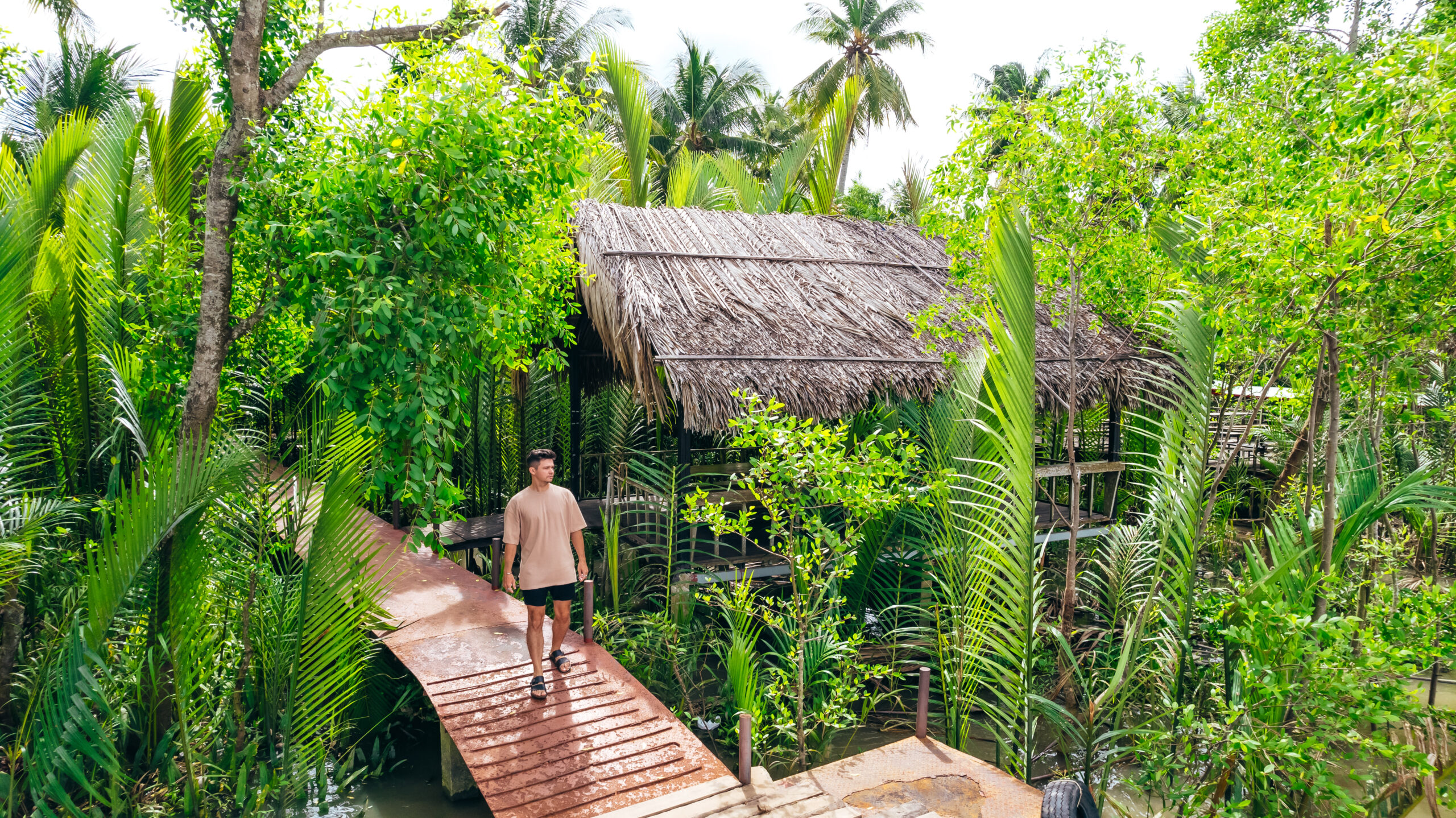 young man walk down pier of Ben Tre over looking straw thatched house, surrounded by greenery