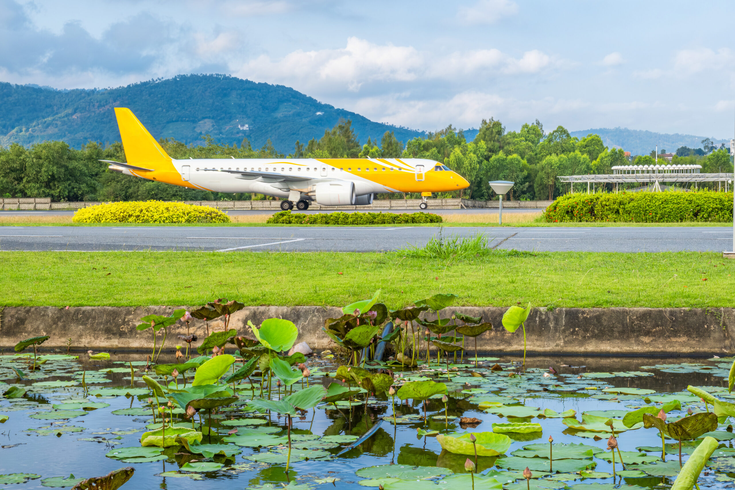 Airplane taxiing at Samui airport with lush green surroundings, lotus pond in foreground and mountains in the background, Thailand